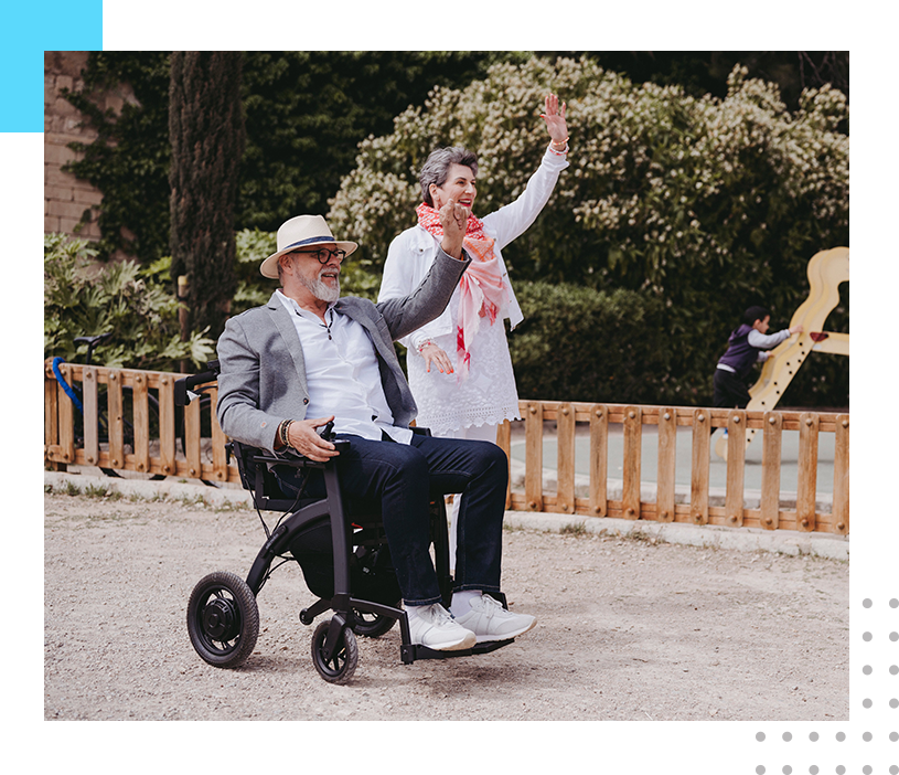 A man in a wheelchair and another person wave at the camera.